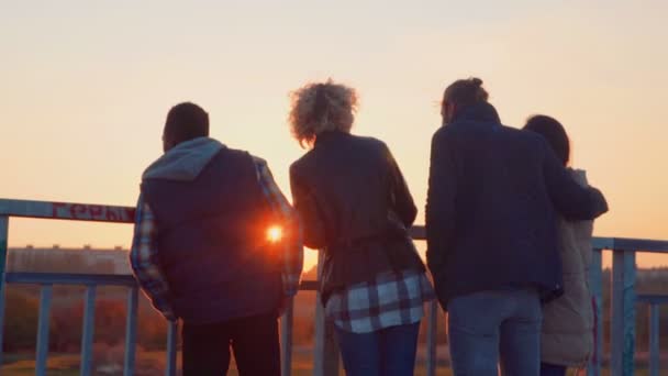 Fyra vänner umgås på bron vid solnedgången. — Stockvideo