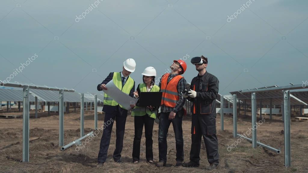 Man controlling the drone in virtual reality goggles while managers discussing the plan of the field with solar panels using computer laptop