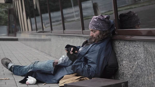 Γενειοφόρος επαιτεία άνθρωπος που κάθεται στο δρόμο με το smartphone. — Φωτογραφία Αρχείου