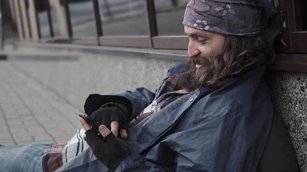 Barbudo mendigo sentado en la calle con smartphone . — Foto de Stock