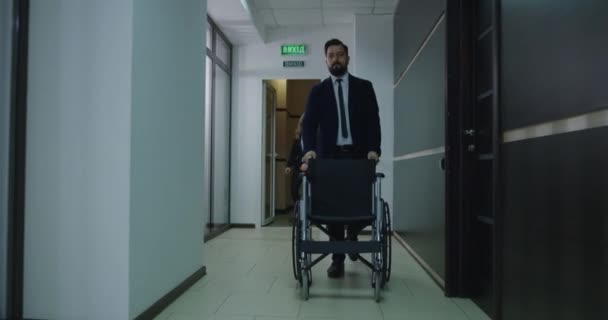 Бизнесмен толкает пустую инвалидную коляску в служебный коридор — стоковое видео