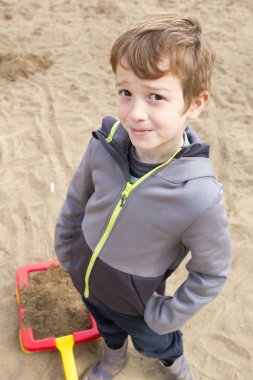 Kızıl saçlı çocuk kameraya bakarak kumla oynuyor.