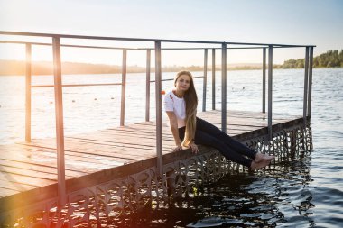Kadın iskelede oturuyor, suya ve günbatımına karşı