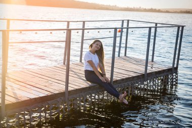 Kadın iskelede oturuyor, suya ve günbatımına karşı
