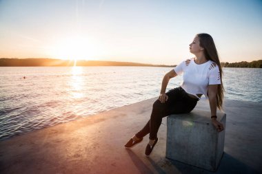 Kadın iskelede oturuyor, suya ve günbatımına karşı