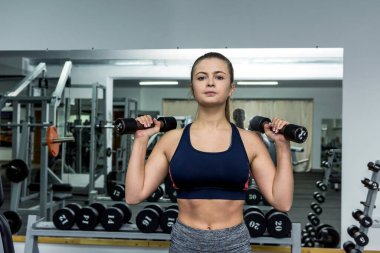 Güzel bir kadın spor salonunda dambıllarla tahta egzersizi yapıyor.