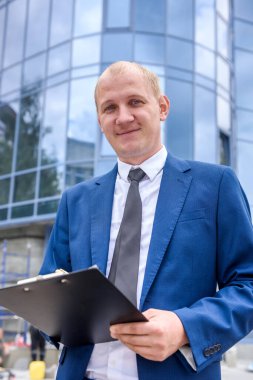 Takım elbiseli gülümseyen adam iş merkezinin önünde poz veriyor.