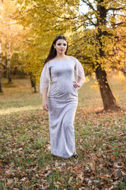 Sonbahar parkında poz veren zarif gece elbiseli bir kadın. Ağaçlardaki güneş ışıkları sarı renklidir.