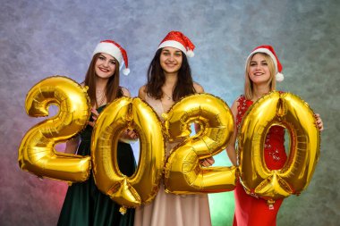 Noel Baba şapkalı üç neşeli kız 2020 numaralı balonlarla parti kutlamalarında poz veriyor.