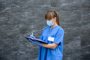 Not defterli bir doktor. Mavi ceketli kadın gri arka planda steteskoplu poz veriyor.