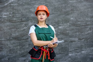 Tulumlu ve kasklı bir kadın elinde not defteri ve gri arka planda kameraya poz veriyor.