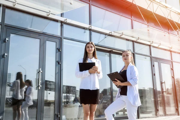 Two young business women with tablet and clipboard posing outside business center