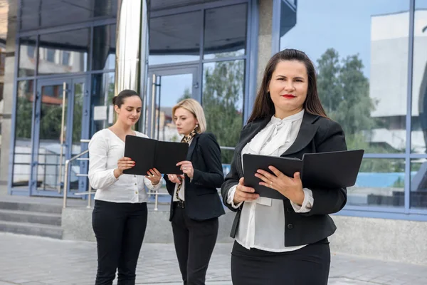 Businesswoman with tablet looking in camera. Her colleagues discussing information in folder