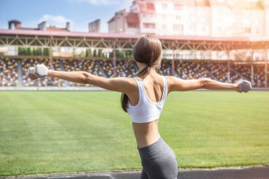 Zayıf kadın sabah egzersizlerini açık havada dambıllarla yapıyor. Spor kıyafetleriyle stadyumda poz veriyor.