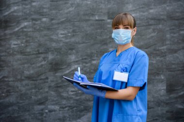 Not defterli bir doktor. Mavi ceketli kadın gri arka planda steteskoplu poz veriyor.