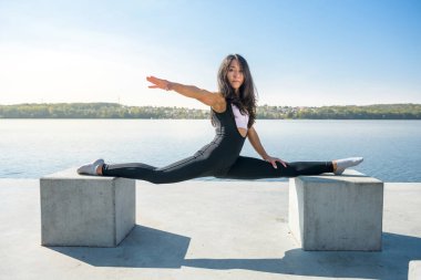 Genç Avrupalı bayan sporcu bacaklarını esnetme egzersizleri yapıyor, dışarıda göl kenarındaki yarıklara oturuyor. Sağlıklı yaşam tarzı