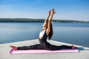 Genç Avrupalı bayan sporcu bacaklarını esnetme egzersizleri yapıyor, dışarıda göl kenarındaki yarıklara oturuyor. Sağlıklı yaşam tarzı