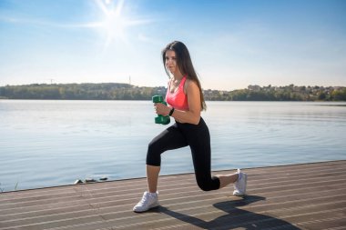 Formda kız, gölün kenarında, elinde dambıllarla egzersiz yapıyor.