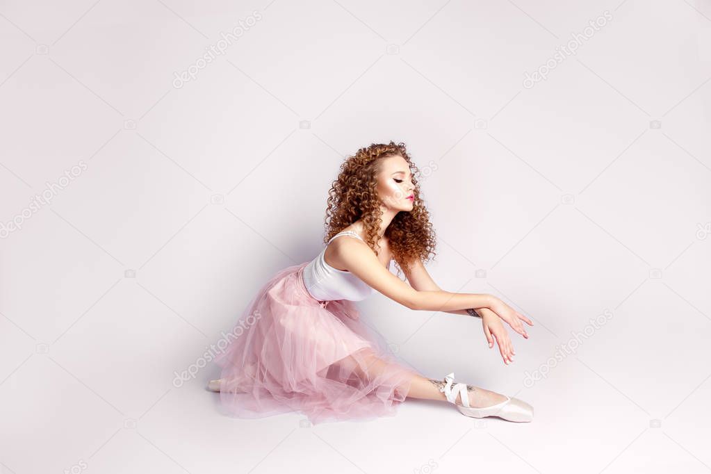 Young curly ballerina in pink dress and ballet shoes posing in studio