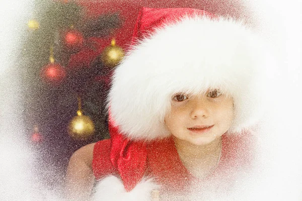 Kind in Weihnachtsmütze mit Weihnachtsbaum am Fenster. Kind und Schneefall. — Stockfoto