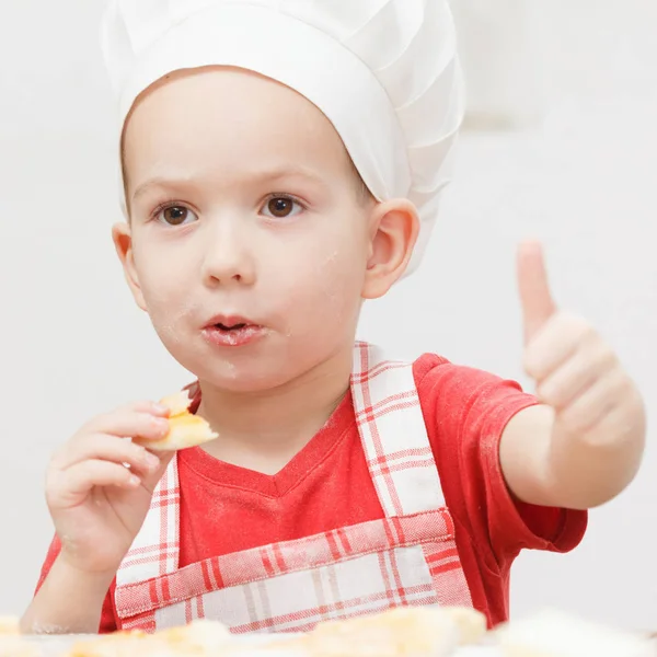 Little cheerful boy chef cooks and eats pizza — Stock Photo, Image