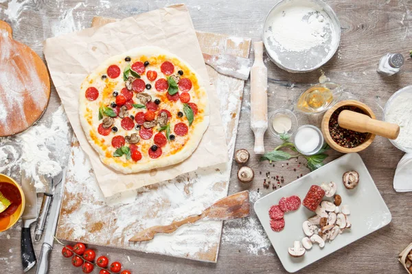 Pizza mit Zutaten, Tomaten, Salami und Pilzen auf hölzerner Tischplatte — Stockfoto