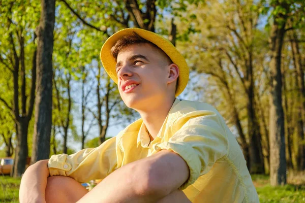 A portrait of a happy teenage boy outside, wearing a yellow shirt and hat against a blue sky, green tree — Stock Photo, Image