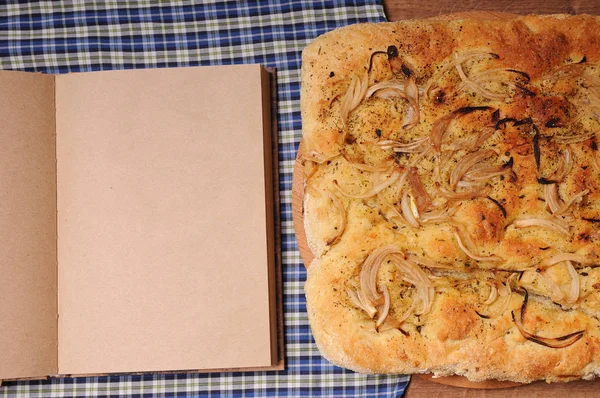 Луковый итальянский хлеб и открытая тетрадь с бесплатным местом для текста. На деревянном столе со скатертью в клетку — стоковое фото