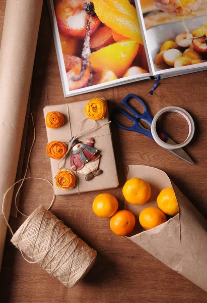 Paquete de regalo de Navidad en estilo kraft. Mandarinas, libro, cuerda de yute, rollo de papel, tijeras, whisky sobre mesa de madera — Foto de Stock