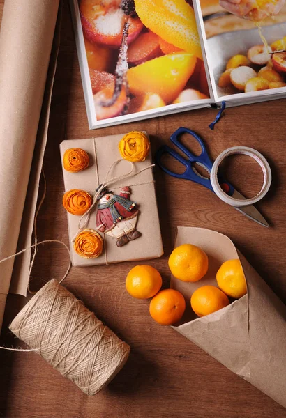 Paquete de regalo de Navidad en estilo kraft. Mandarinas, libro, cuerda de yute, rollo de papel, tijeras, whisky sobre mesa de madera — Foto de Stock