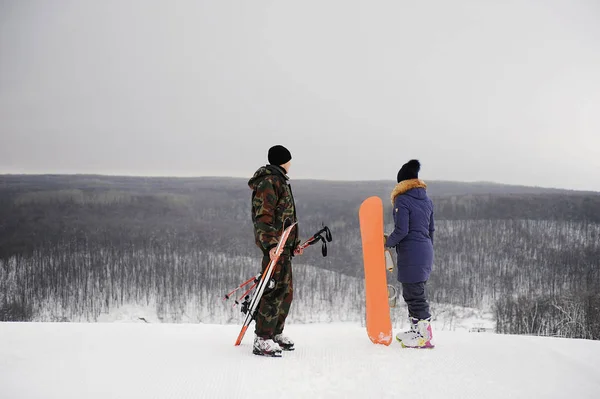 Człowiek i dziewczyna z snowboardowe i narty, stojąc na szczycie monochromatyczne tła z zimowego krajobrazu — Zdjęcie stockowe