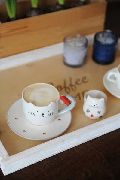 Kávéfőző cappuccino-aranyos csésze alakú macska. Egy fa tálcán. Gyertyák a háttérben — Stock Fotó