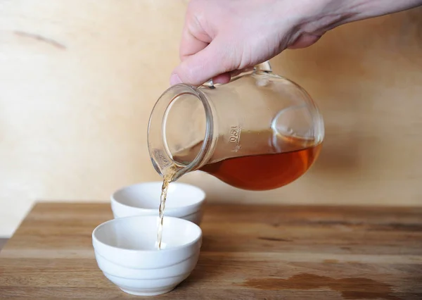 Hand gieten thee of koffie uit een karaf van glas op de kom van porselein. Houten achtergrond. — Stockfoto