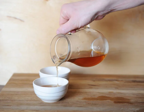 Ręka nalewania kawy lub herbaty z karafki szkła na miskę porcelany. Podłoże drewniane. — Zdjęcie stockowe