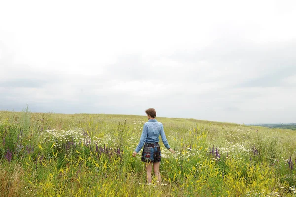 Menina em uma camisa de ganga fica com as costas no fundo de um prado selvagem de verão com flores — Fotografia de Stock