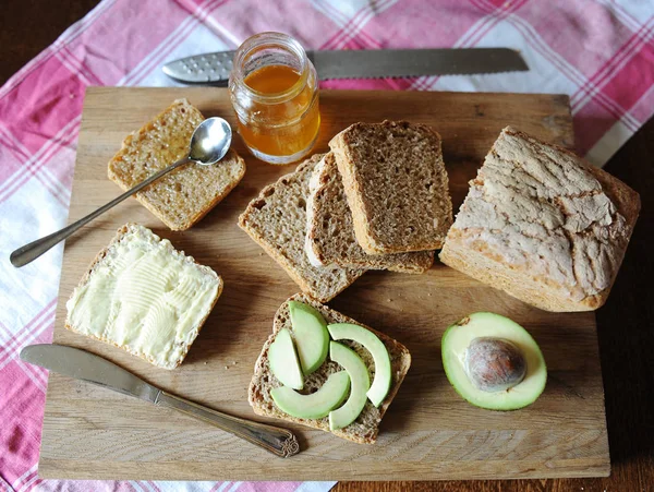 Desayuno saludable con pan fresco, aguacate, mermelada de miel y mantequilla en una tabla de madera sobre mantel a cuadros —  Fotos de Stock