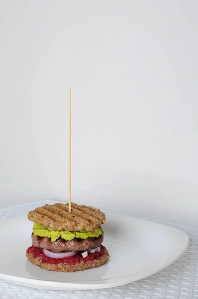 Hamburguesa con empanadas de ternera, pan de trigo sarraceno, salsa de guacamole, cebollas frescas y cerezas. En un plato blanco sobre un fondo blanco en la parte izquierda del marco —  Fotos de Stock