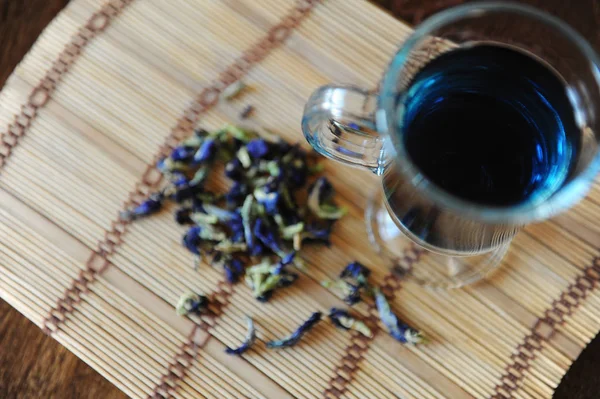 Ancla de té tailandés azul en taza de vidrio en estera de bambú en mesa de madera, vista superior. Colocador de flores del clítoris junto a copa — Foto de Stock