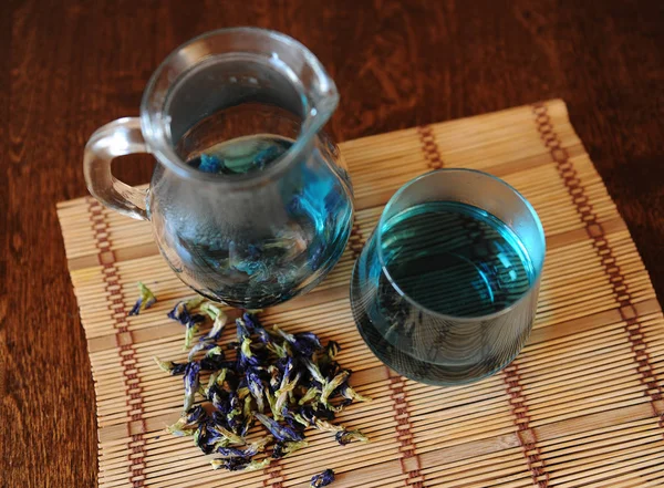 Кувшин и стакан с голубым тайским чаем анчан на бамбуковом коврике на деревянном столе. Размещение цветов для пивоварения, вид сверху — стоковое фото