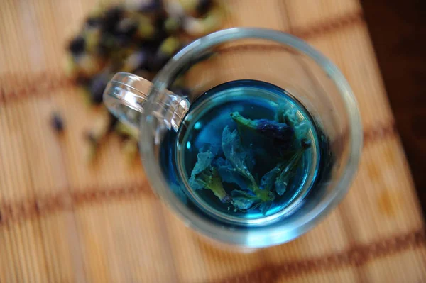 Голубой тайский чай анчан в стеклянной чашке на коврике на деревянном столе, вид сверху. Гипсокартон из цветов клитории рядом с чашкой — стоковое фото