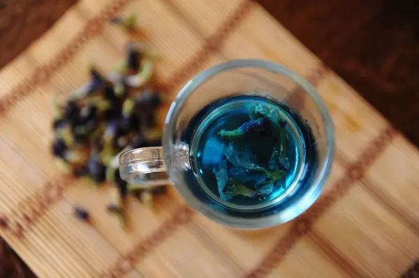 Ancla de té tailandés azul en taza de vidrio en estera de bambú en mesa de madera, vista superior. Colocador de flores del clítoris junto a copa — Foto de Stock