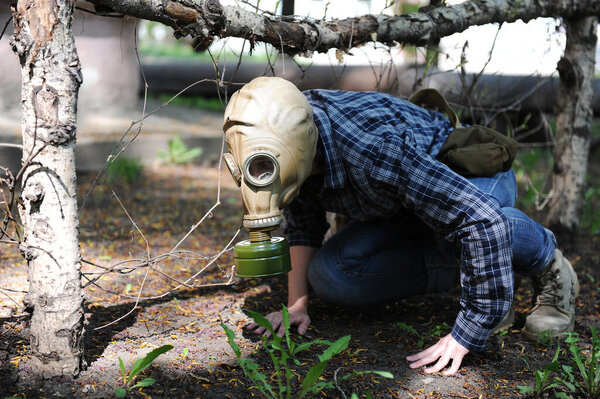 A man in a gas mask creeps on the ground with dry trees and spring greens. Environmental pollution