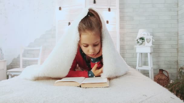 Портрет милой школьницы, читающей книгу в пижаме, покрытую белым шерстяным одеялом . — стоковое видео