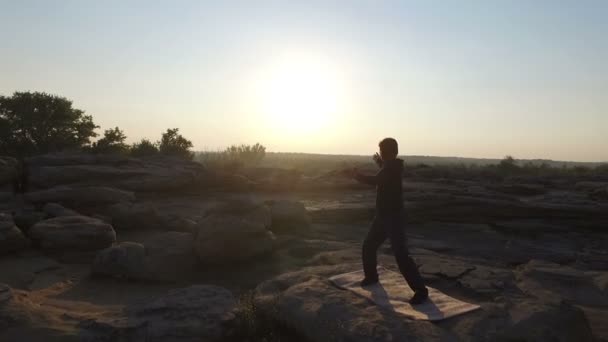 Un homme engagé dans la gymnastique chinoise, sur les rochers au coucher du soleil. Aérien . — Video