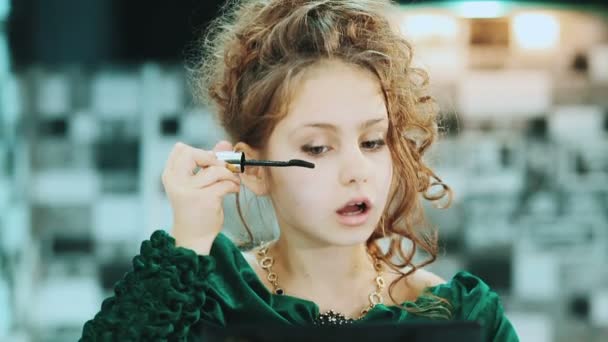 Linda niña en un vestido verde pestañas de color sentado cerca de un espejo . — Vídeo de stock