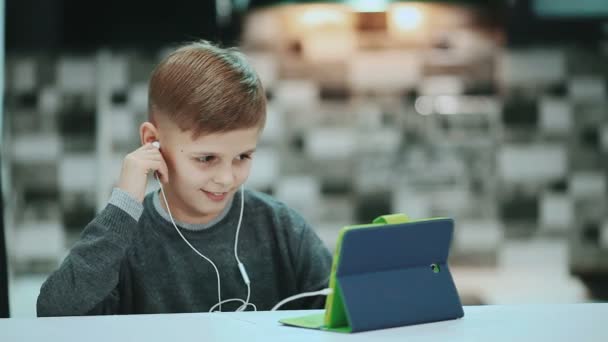 Маленький мальчик в наушниках использует цифровой планшет, сидит дома и слушает музыку — стоковое видео