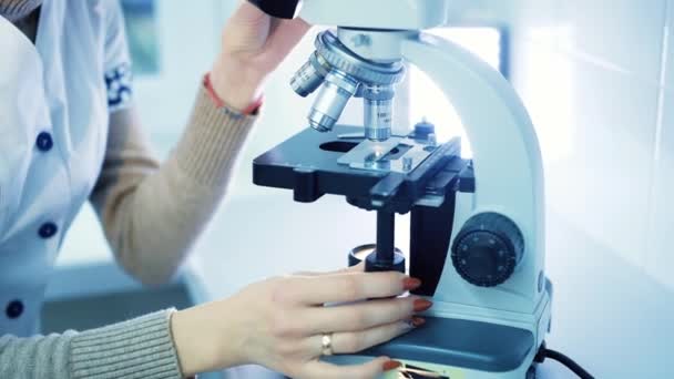 Asistente profesional de laboratorio femenino en una capa blanca trabaja detrás de un microscopio . — Vídeo de stock