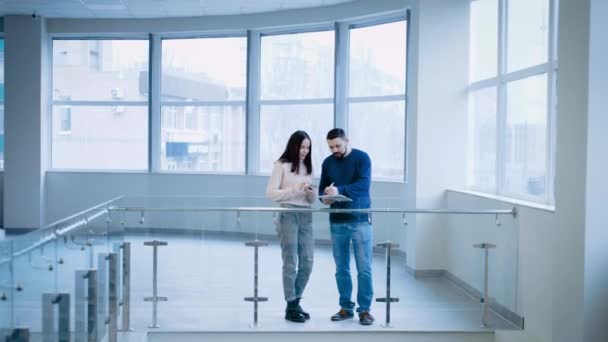 Colleagues stand in a spacious office building and discuss the project, actively writing notes into the workbook. — Stockvideo