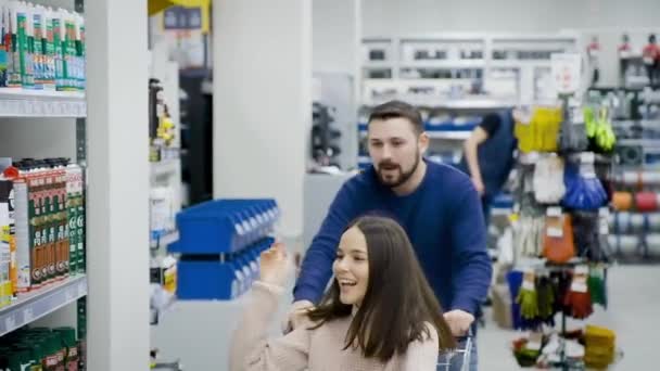 Guy rolt zijn aantrekkelijke jonge vriendin in de trolley in de supermarkt. — Stockvideo