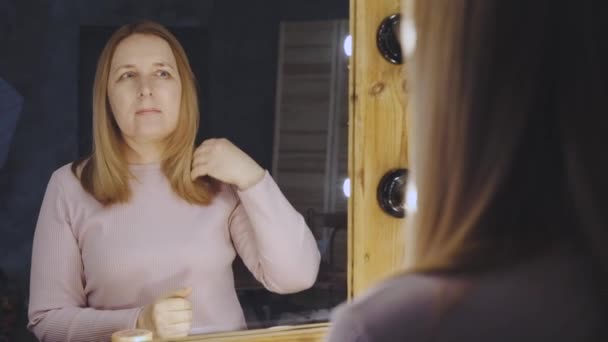 Блондинка зріла жінка налаштовує пряме волосся, стоячи біля дзеркала — стокове відео